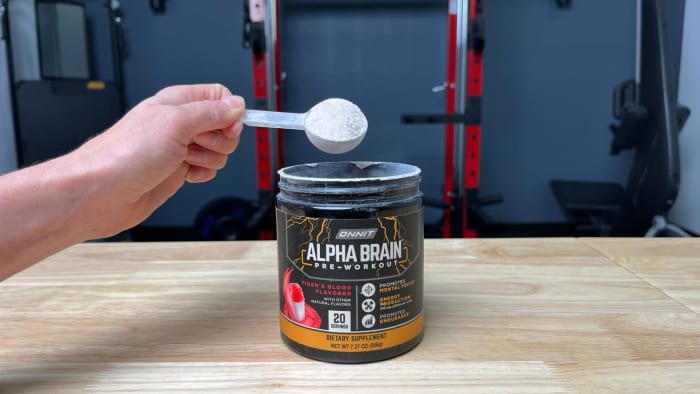 A person in a gym holding a scoop of Onnit Alpha Brain Pre-Workout powder in Tiger's Blood flavor